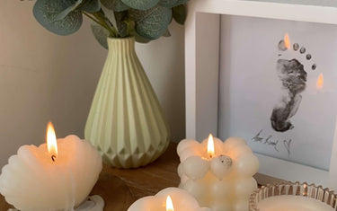 Burning candles on tables, with framed picture of a child's foot and plant in a vase in the background