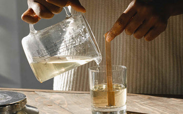 Woman pouring melted candle wax into a clear glass candle jar with a wooden wick
