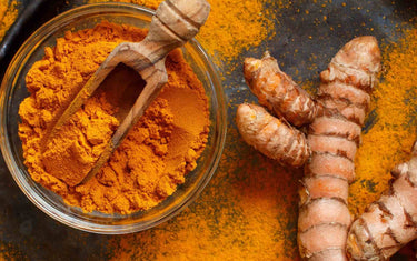 Glass bowl with powdered turmeric and a wooden spoon next to whole turmeric root