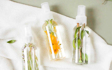 Three clear spray bottles with white lids resting on a towel. Spray bottles contain clear liquid and various floral and leaf botanicals.
