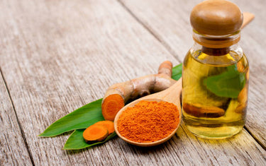 Small glass vial of turmeric oil next to a wooden spoonful of turmeric powder and a thumb sized piece of turmeric root