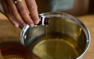 Womans hand pouring oil from an amber glass essential oil bottle into a pan with melted candle wax in it