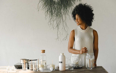 Woman looking to the side with various equipment for making candles on a wooden table. Just above her head is a dangling plant