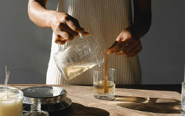 Woman pouring melted wax into a clear glass candle container with a wooden wick in it