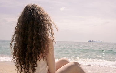 Woman with long curly hair sits on beach and looks at sea