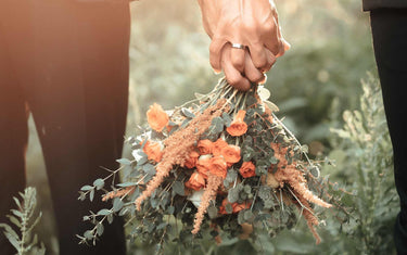 Couple holding hands and a bouquet with greenery behind them