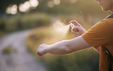 Man applying repellent spray to his arm. He's wearing an orange t shirt and the background shows a blurred pathway surrounded with grass and probably trees.