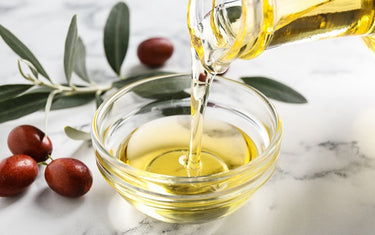 Jojoba oil being poured into a small glass ramekin with some parts of the jojoba plant in the near background
