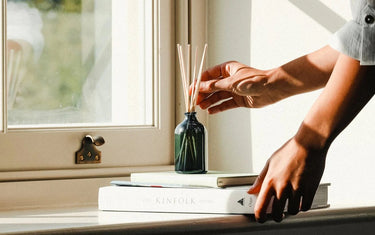 Womans hand adjusting a reed diffuser. The diffuser is placed on a couple of books next to a window with sunshine coming through
