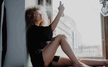 Woman by a window spraying mist on her face.