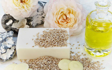 Bottle of sesame oil next to sesame seeds, soap, and flowers.