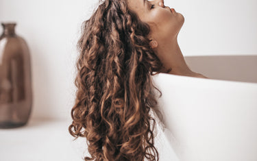 Women with long curly hair rests her head on the side of a bath